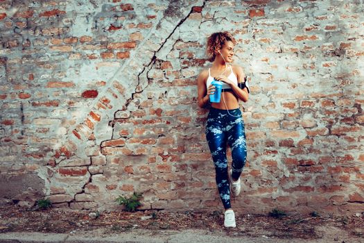 Young muscular woman with headphones standing up against the wall and resting after jogging.