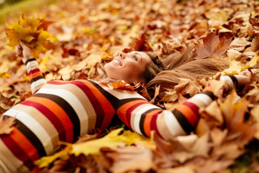 Beautiful young woman in sunny forest in autumn colors. She is lying on the ground covered with leaves and enjoying. 