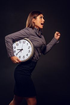 Stressed businesswoman in a hurry running with clock under her arm because she late. Dark background.