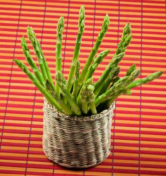 Bunch of Fresh Raw Asparagus Sprouts in Wicker Box closeup on Orange Straw Mat background