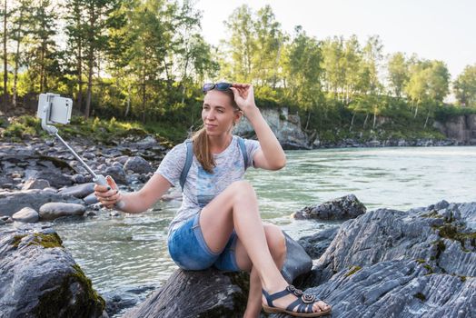 Woman taking selfie on mobile phone with stick on mountain river background