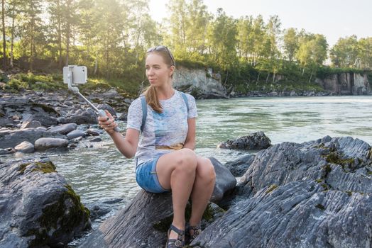 Woman taking selfie on mobile phone with stick on mountain river background