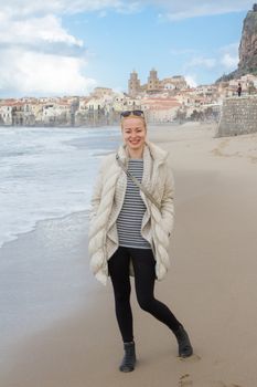 Female tourist posing for photo on empty beach of Cefalu in out of touristic season in winter time, Sicily, south Italy.