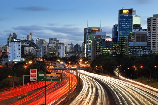 Traffic lights streak along the expressway into and out of the city at dusk.  Sydney icons, like Centrepoint Tower and Sydney Harbour Bridge in view as well as many building of the CBD and North Sydney.