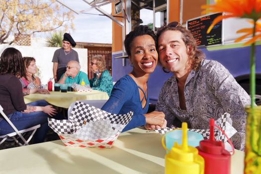 Handsome diverse couple smiles at camera while seated at an outdoor table beside a food truck