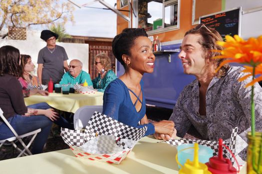Diverse couple smiles at one another while seated beside a food truck