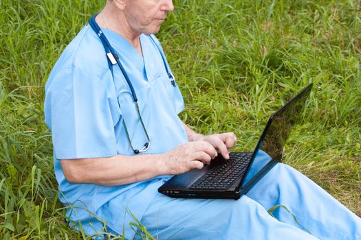 Doctor concept. A man with a stethoscope and in a medica gown is holding a laptop on his lap. Sitting on the grass. Photo for your design on the background of nature. Horizontal sheet orientation