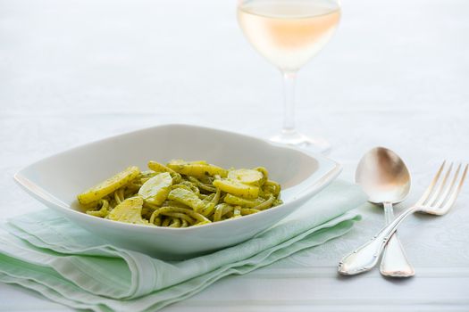 Linguine pasta with pesto genovese and potatoes over a table with cutlery and white wine glass