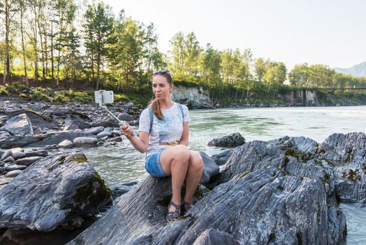 Woman taking selfie on mobile phone with stick on mountain river background