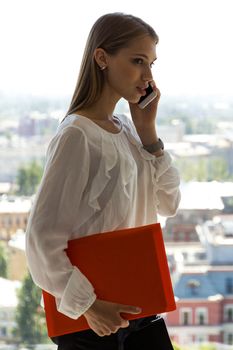 Young businesswoman with folder talking on mobile phone with client standing near office window with view of city