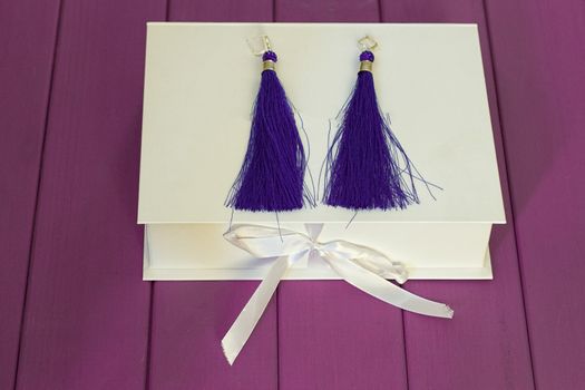 Handmade blue threaded earrings and a white box on a purple wooden background