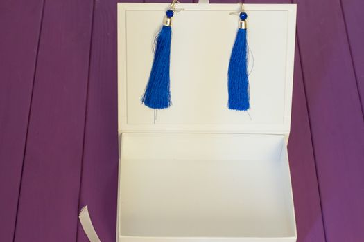 Handmade blue threaded earrings and a white box on a purple wooden background