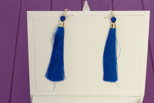 Handmade blue threaded earrings and a white box on a purple wooden background