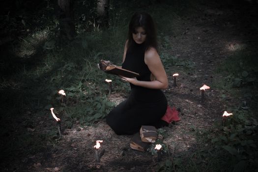 Halloween concept. Fairy in black. In the witchy circle. With a book of spells in hand. Looks up. Among woods and trees. Sideways