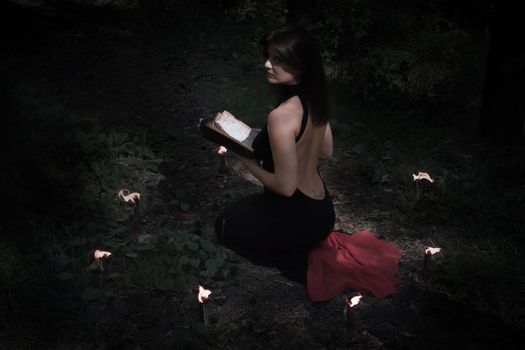 Halloween concept. Fairy in black. In the witchy circle. With a book of spells in hand. Looks up. Among woods and trees. With a beautiful back