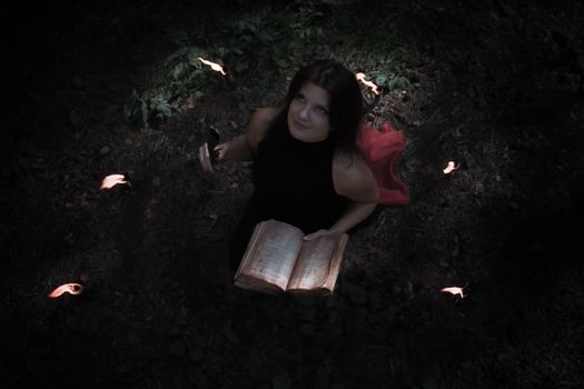 Halloween concept. Witch in black. In the witchy circle. With a satanic book in his hands. Looks up. Among woods and trees. Lighted candles of fire. Witchcraft and magic.