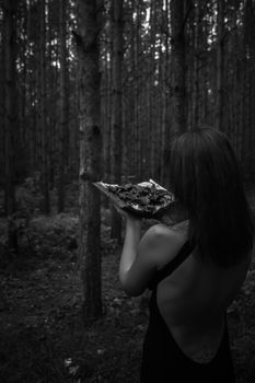 Halloween concept. A witch in black with a witch satanic book. Among the forest of the trees. Photo for your design. Vertical sheet orientation