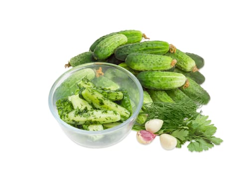 Lightly salted cucumbers in the glass bowl against of a pile of the freshly picked out cucumbers, parsley, dill and garlic on a white background
