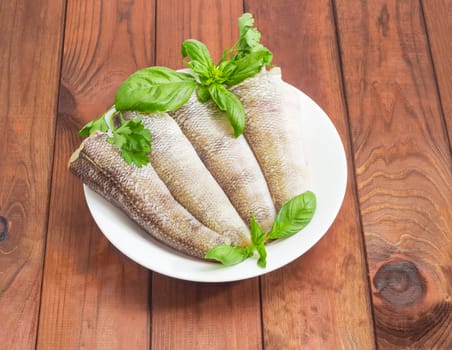 Uncooked carcasses of the notothenia fish without of a heads and tails and with peeled scales and prepared for cooking, twigs of basil and parsley on the white dish on a dark wooden surface
