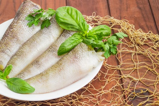 Fragment of a white dish with carcasses of the notothenia fish without of a heads and tails and with peeled scales, basil and parsley twigs on a dark wooden surface with fishing net closeup
