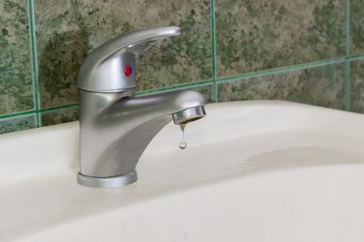 Drop of water from the single handle mixer tap mounted on a wash basin on background of a wall with green tiles
