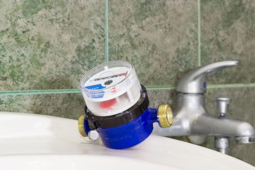 Not connected residential water meter for consumption measuring of a cold water on a wash basin on background of a handle mixer tap and wall with green tiles
