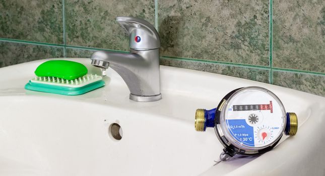 Not connected residential water meter for consumption measuring of a cold water on a wash basin on background of a water dripping from handle mixer tap and wall with green tiles
