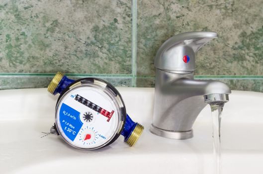 Not connected meter for consumption measuring of a cold water on a wash basin beside mounted handle mixer tap and water flowing from him on background of a wall with green tiles
