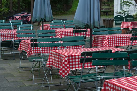 Empty beer tables with red white tablecloth and green wooden chairs in a public beer garden.
