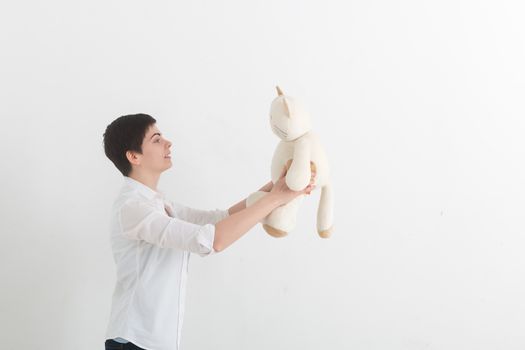 Strange young woman with short hair in white shirt talking to her toy plush cat on light grey background.
