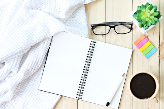 Business, weekend, holiday or good morning concept : Flat lay or top view of white knitted blanket, open blank notebook paper, coffee cup and eyeglasses on wooden background