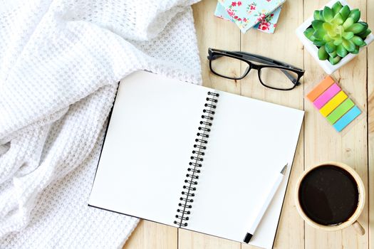 Business, weekend, holiday or good morning concept : Flat lay or top view of white knitted blanket, open blank notebook paper, coffee cup and eyeglasses on wooden background