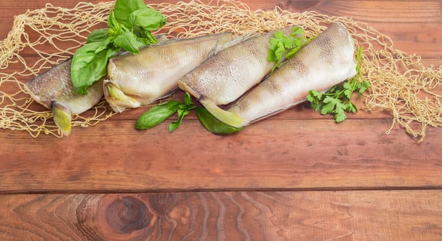 Several uncooked carcasses of the notothenia fish without od head and tail and twigs of basil and parsley on the fishing net on a dark wooden surface
