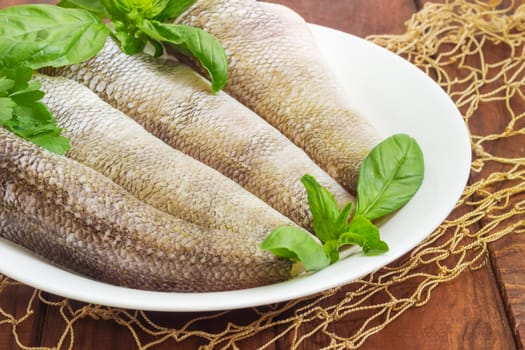 Fragment of a white dish with carcasses of the notothenia fish without of a heads and tails and with peeled scales, basil and parsley twigs on a dark wooden surface with fishing net closeup
