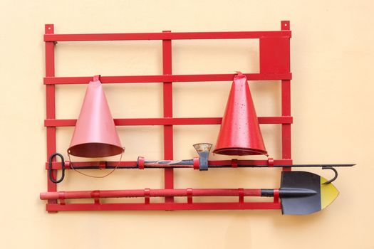 Red fire stand with fire extinguishing tools: conical fire buckets, fire ax, hook and spade hanging on a yellow wall  
