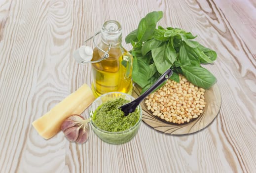 Sauce basil pesto in the small glass bowl with small black ceramic spoon on a background of ingredients for its preparation on a wooden surface
