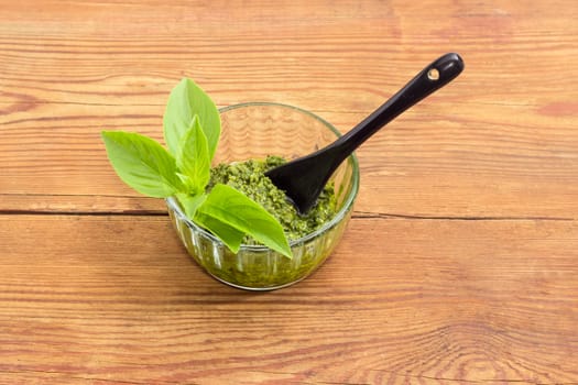 Sauce pesto in the small glass bowl with small black ceramic spoon and decorated with green basil twig on a surface of an old wooden planks
