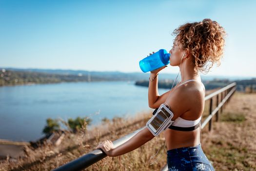 Young urban runner woman with headphones resting after jogging and drinking water on the city rooftop.