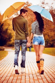Beautiful lovely couple with umbrellas enjoying while walking in the rain through the park in autumn colors. Rear view.