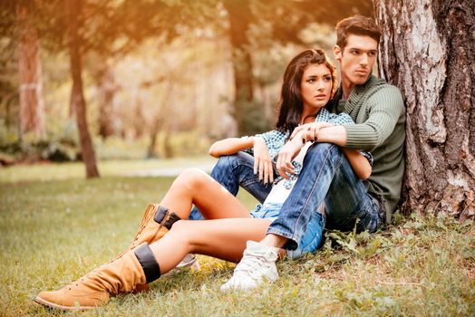 Beautiful lovely couple sitting on the grass next to the tree and enjoying in sunny park in autumn colors.