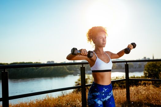 Young fitness woman doing workout with dumbbell by river on the city rooftop.