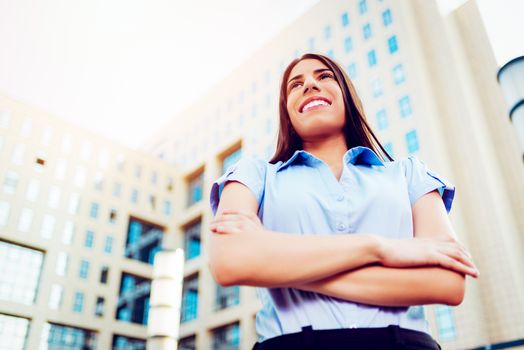 Young smiling businesswoman standing with crossed arms in office district and looking away. 