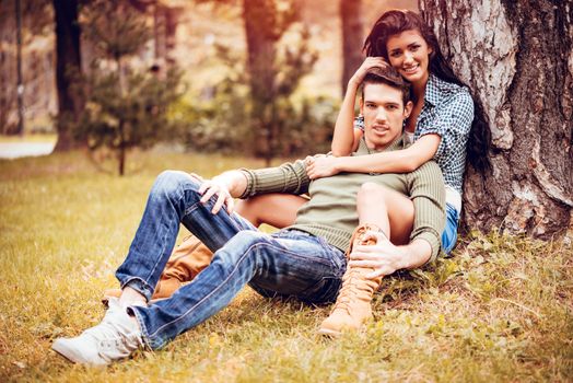 Beautiful lovely couple sitting on the grass next to the tree and enjoying in sunny park in autumn colors. Looking at camera.