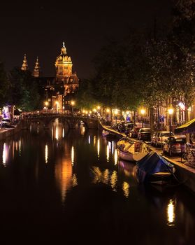 View of a church and a canal in Amsterdam, Netherlands at night. The Basilica of Saint Nicholas (Sint-Nicolaasbasiliek) with the reflection in the canal