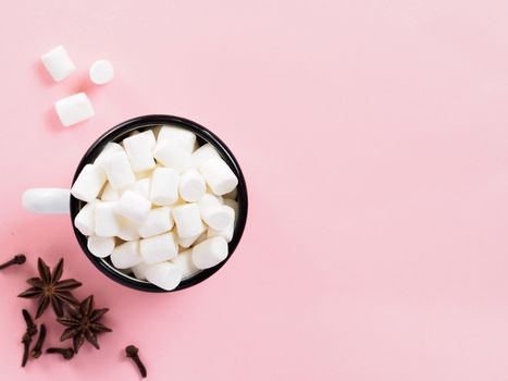 Marshmallows in cup and winter spices on pink background with copyspace. Flat lay or top view. Background or texture of colorful mini marshmallows. Winter food background concept.