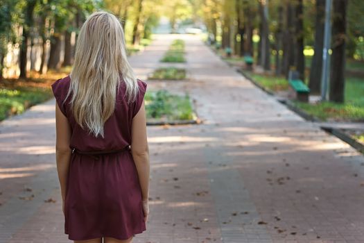 Blonde girl with long hair in burgundy dress standing in the park with her back to the camera