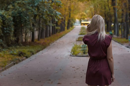 Blonde girl with long hair in burgundy dress standing in the park with her back to the camera