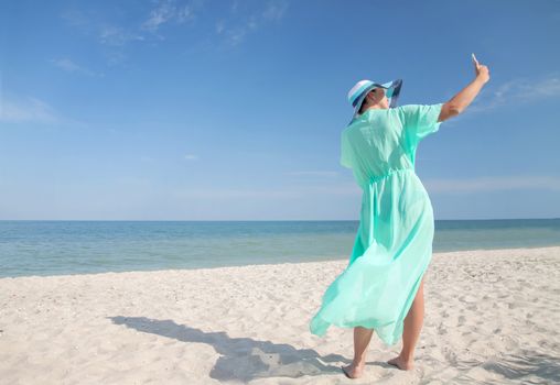 Beautiful young woman in a turquoise dress and a wide hat doing salvi on the phone on the beach, against the backdrop of the sea and sky.