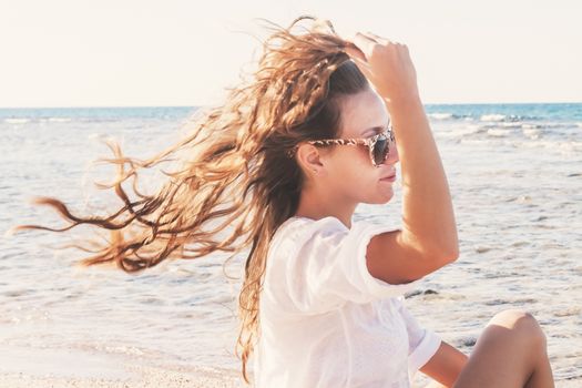 A young woman in sunglasses holds a hand to her hair fluttering in the wind near the sea. The concept of a free way of life.