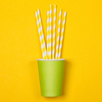 Multicolored cocktail tubes in green paper disposable cup on a yellow background